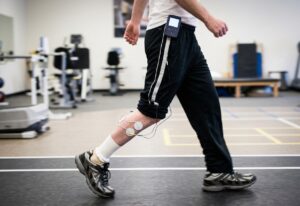 A person wearing an electrical stimulation device and electrodes on their lower leg, with wires connecting to a waist-mounted control unit, while walking in a rehabilitation gym.