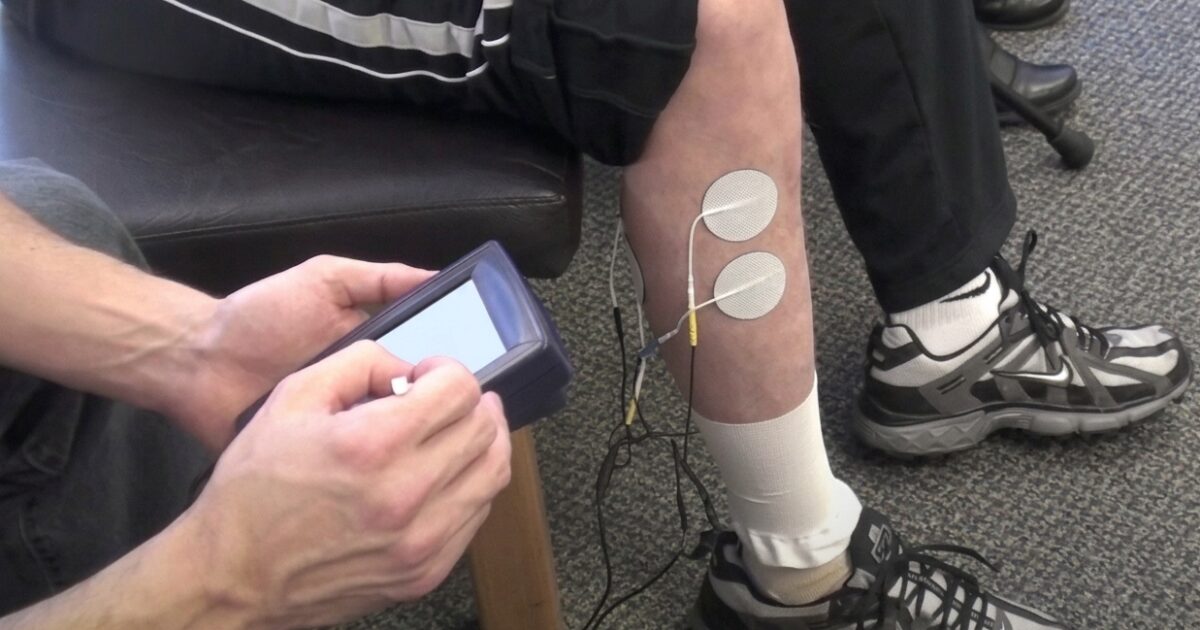 a person having electrodes and wires attached to their leg, with another person holding a control device nearby.