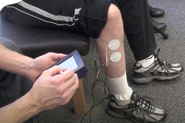 a person having electrodes and wires attached to their leg, with another person holding a control device nearby.